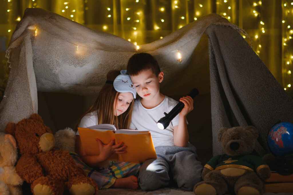 Two siblings reading a book by flashlight inside a cozy blanket fort decorated with twinkle lights.